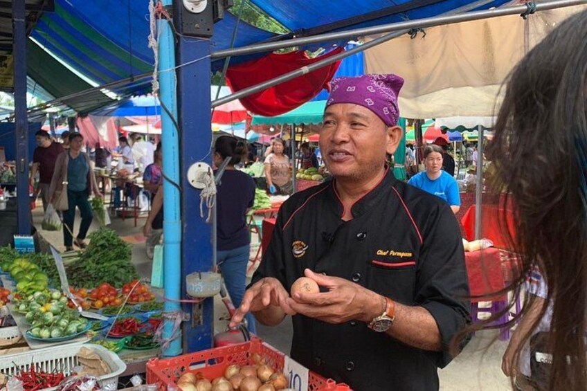 Escena de mercado con un vendedor con uniforme de chef negro y pañuelo morado, rodeado de productos frescos y un toldo azul. 2