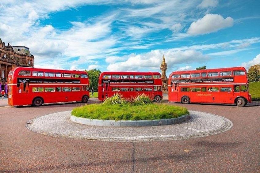 Drie rode dubbeldekbussen geparkeerd in een cirkelvormig gebied met een grasveld in het midden, omgeven door een verharde weg en een historisch gebouw op de achtergrond. 2
