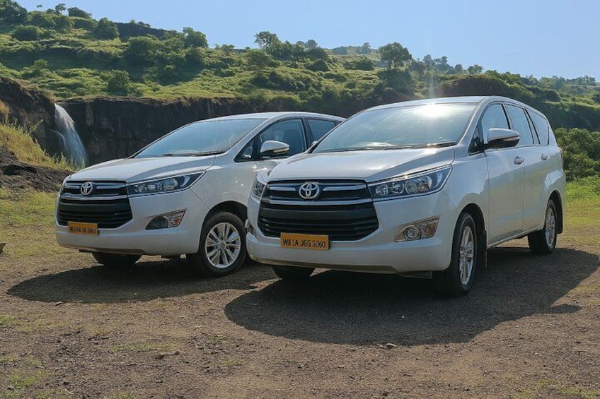 Två vita Toyota-terrängbilar står parkerade på en grusväg, med ett vattenfall och frodig grönska i bakgrunden. 1