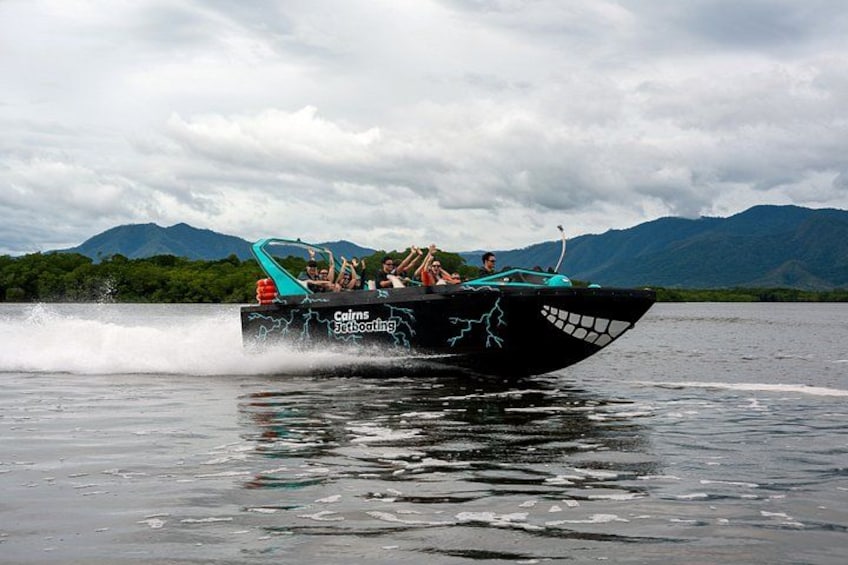 Ein Schnellboot mit der Beschriftung 'Cairns Jetboating' an der Seite, das über ein Gewässer fährt, wobei sich Berge im Hintergrund abzeichnen. 1