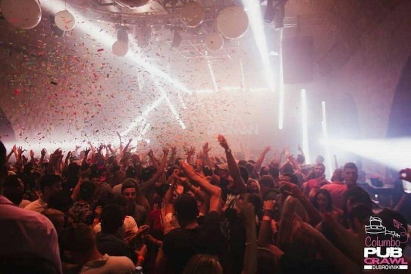 A crowded indoor event with people raising their hands, confetti falling, and stage lighting. 3