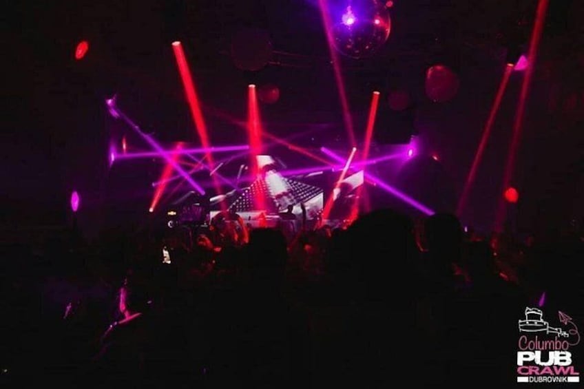 A club interior with a DJ booth, red and purple lighting, and a crowd. 2