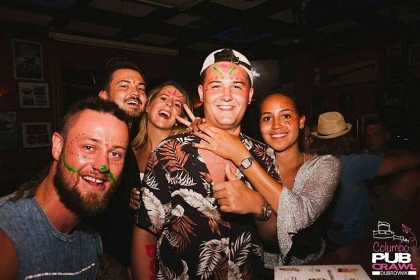 A group of people indoors, some with face paint, in a social setting with a visible logo for 'Pub Crawl Dubrovnik'. 2