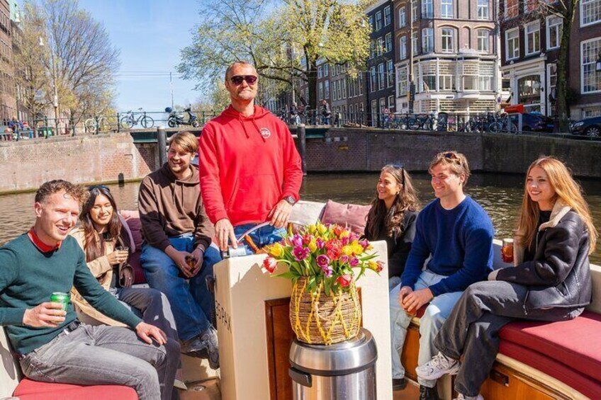 Un grupo de personas en un bote con una mesa en el centro, un canal y edificios al fondo. 2