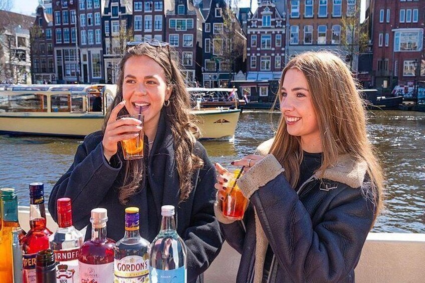 Dos personas brindando con bebidas en una mesa con varias botellas frente a un canal con barcos y edificios históricos al fondo. 3