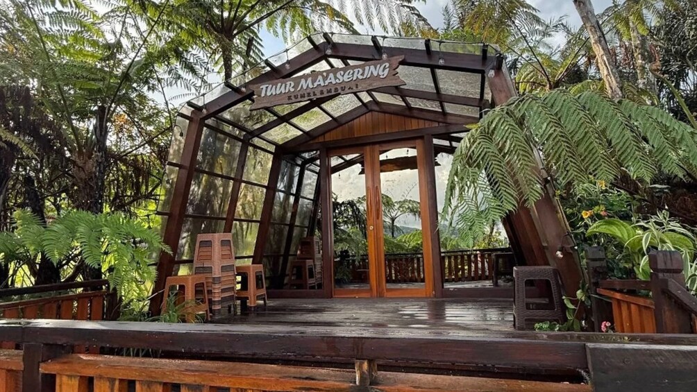 A wooden structure with a sign reading 'Tur Maasering Kumelembual' surrounded by lush greenery. 2