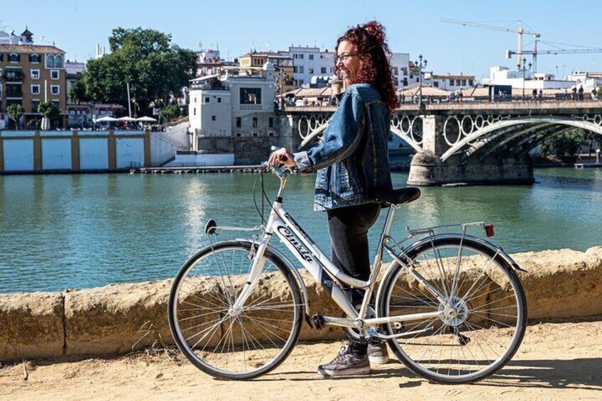 A person standing next to a white bicycle with a basket, near a river with a bridge in the background. 3