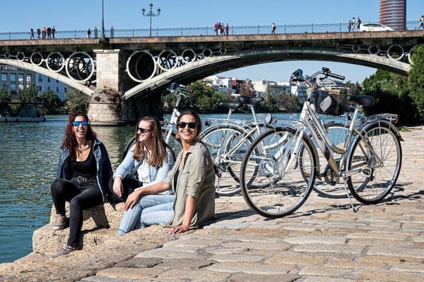Three people sitting on a stone ledge by a river, with bicycles parked behind them and a bridge in the background. 1