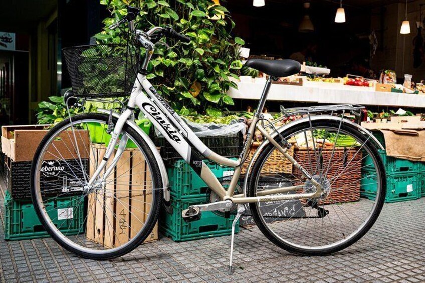 A white bicycle with a basket and a seat, parked on a brick surface next to crates and a market stall. 4