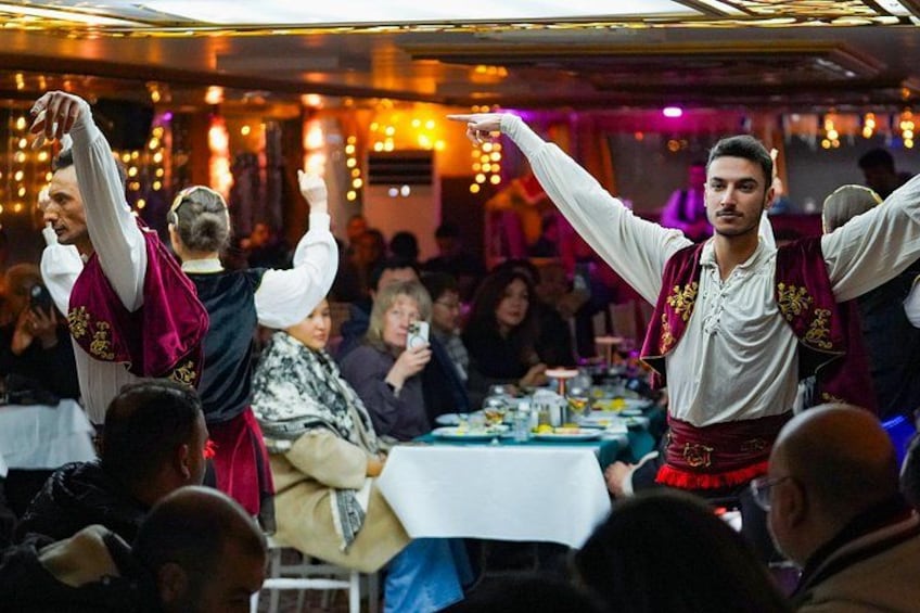 Une personne vêtue d’une chemise blanche et d’un gilet rouge, les bras levés, peut-être en train de danser ou de célébrer, dans un lieu intérieur bondé, avec des tables et d’autres personnes. 2