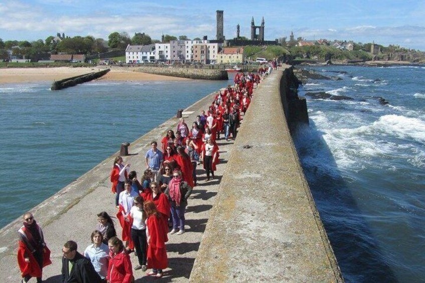 A long pier with people in red attire, a body of water, and a distant building with towers. 4