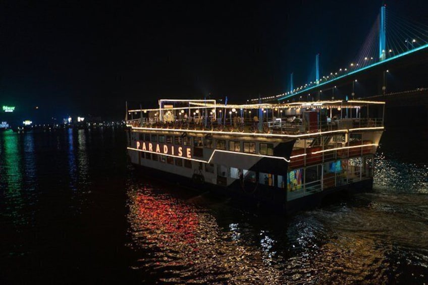 A brightly lit riverboat named 'PARADISE' docked at night with a bridge in the background. 2