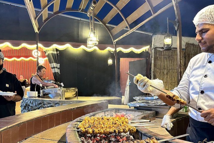 Un puesto de comida con brochetas a la parrilla, un cocinero con uniforme blanco y un escenario con cortina roja al fondo. 2