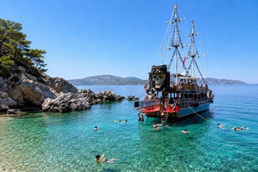 A clear blue sea with a large ship anchored near rocky shores, people swimming, and a mountainous landscape in the background. 2