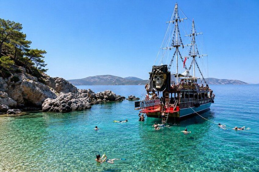 A clear blue sea with a large ship anchored near rocky shores, people swimming, and a mountainous landscape in the background. 4