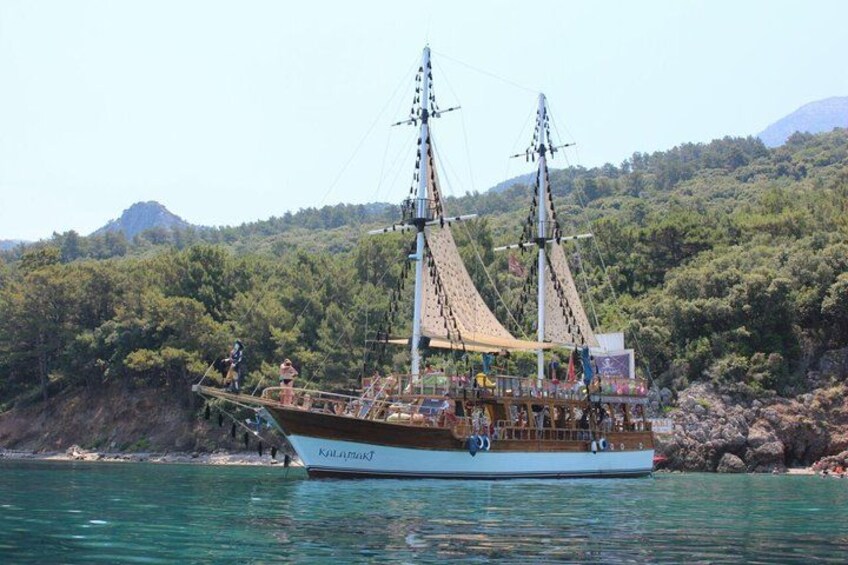 A tall ship with multiple masts and sails docked near a rocky shoreline with dense green trees in the background. 5