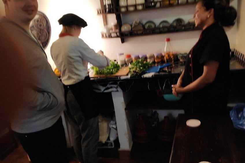 A kitchen with people preparing food, a counter with various ingredients, and a shelf with dishes. 3