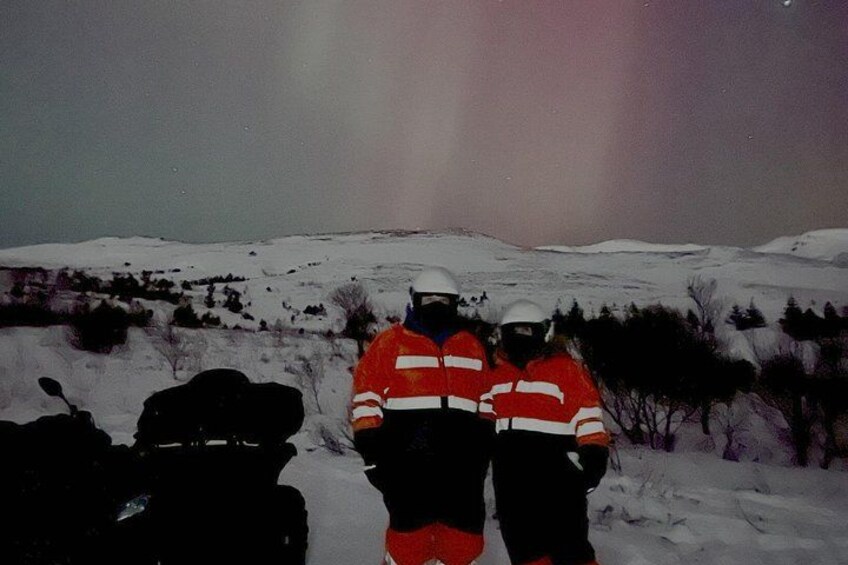 Two people in red and white jackets standing in a snowy landscape with a vehicle in the foreground. 3