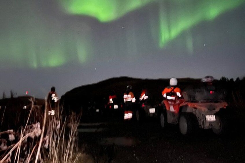 A group of off-road vehicles with drivers wearing helmets and reflective clothing, parked on a dirt road under a night sky with green aurora borealis. 2