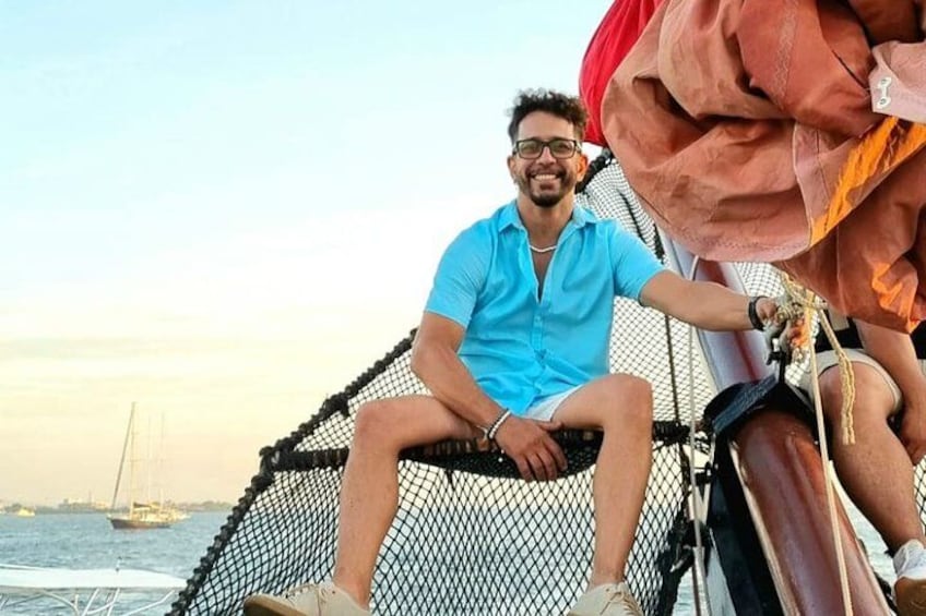 Une personne assise sur un bateau doté d’une rambarde en filet, avec un voilier en arrière-plan et une étendue d’eau. 5