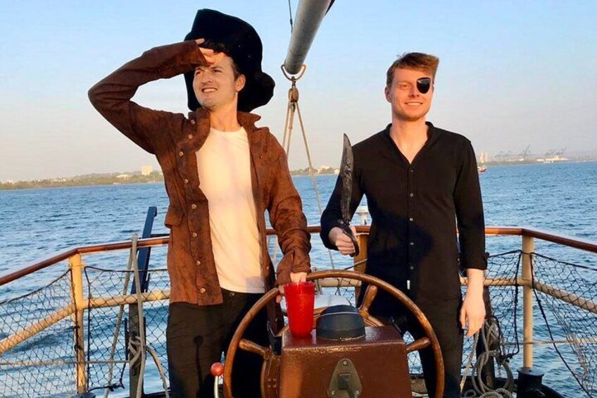 Two people on a boat with a steering wheel, a netted railing, and a body of water in the background. 3
