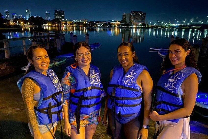 Four people wearing blue life vests by a waterfront with city buildings in the background. 5