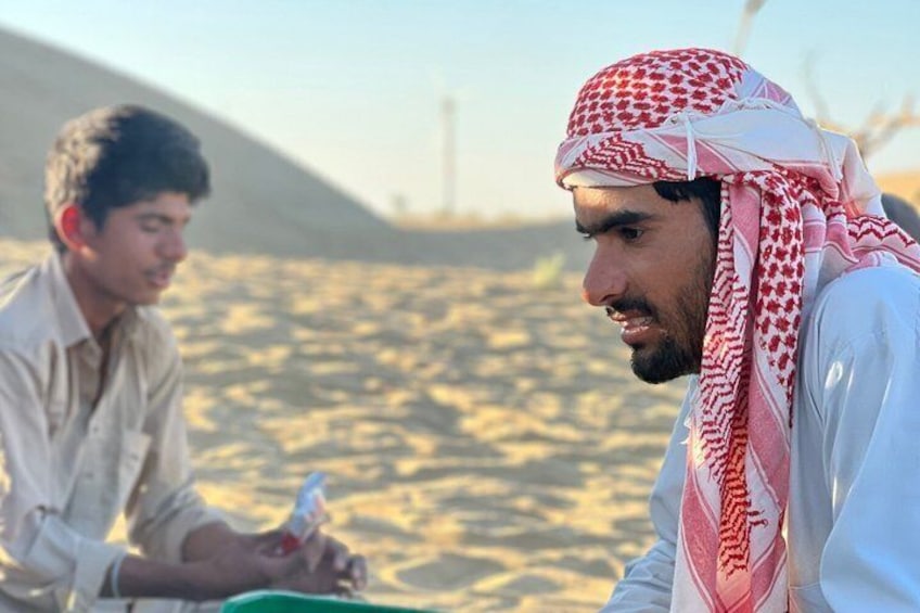 Two men in traditional attire sitting on a sandy beach with a dune and a tower in the background. 1