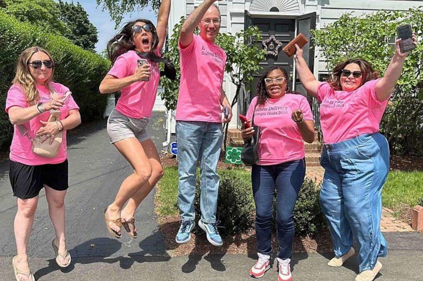 A group of people in pink shirts, some jumping, others holding phones, in a residential area with a house and greenery in the background. 3