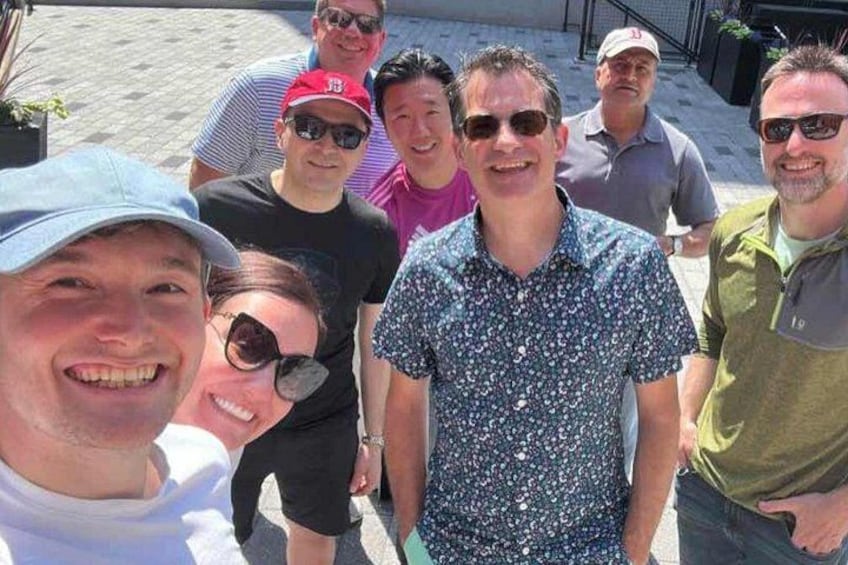 A group of people standing outdoors, some wearing hats and sunglasses, with a paved ground and a fence in the background. 2