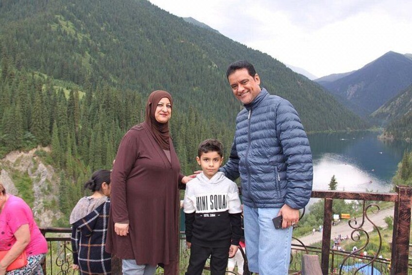 A family of three at a lakeside viewpoint with mountains in the background. 4
