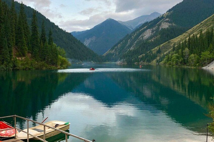 A calm lake surrounded by mountains, a small boat docked on the shore, and a red boat in the water. 2
