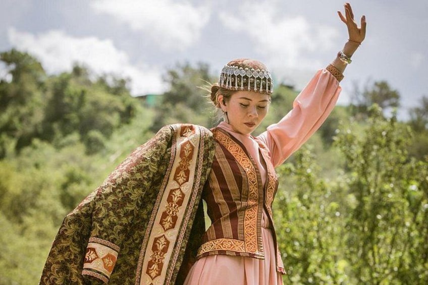 Una persona vestida con traje tradicional, con un chal con estampado y un tocado, de pie al aire libre con follaje de fondo. 5
