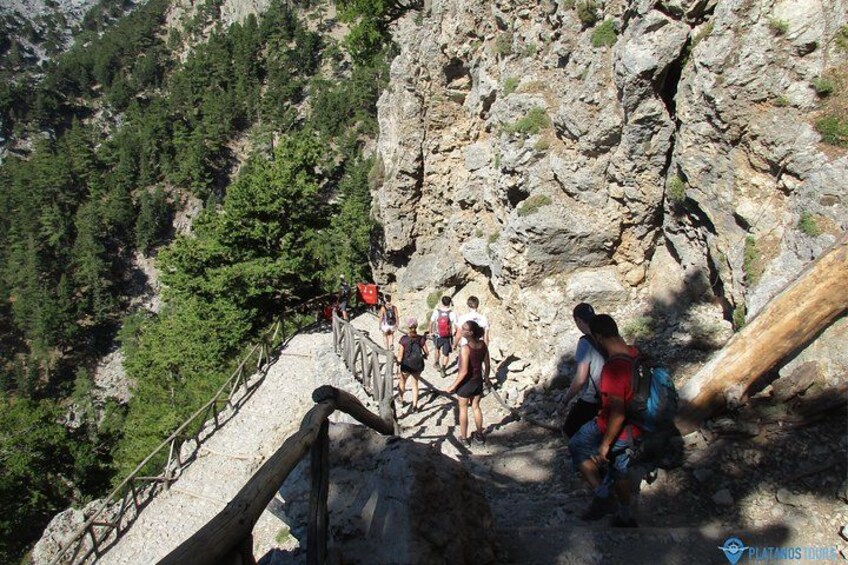 Hikers on a rocky trail with a wooden bridge and forested mountains in the background. 3