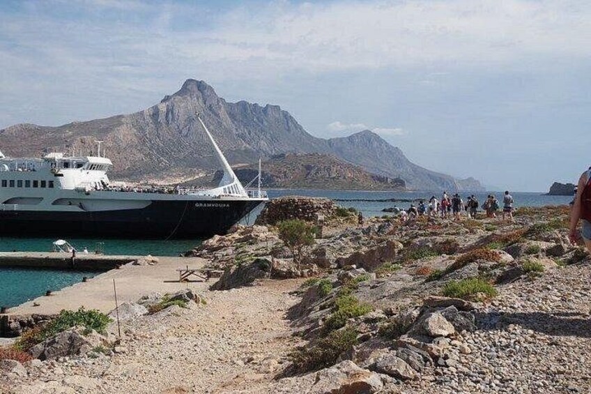 Una costa rocciosa con una barca ormeggiata al molo, uno sfondo montuoso e un gruppo di persone che camminano sulla spiaggia. 2