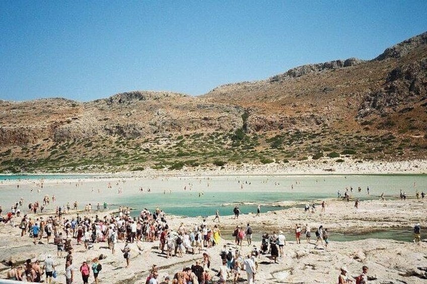 Una playa concurrida con gente caminando y de pie, rodeada de colinas rocosas y agua azul cristalina. 2