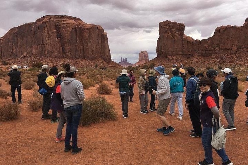 A group of people standing on a dirt path in a desert landscape with large red rock formations in the background. 5