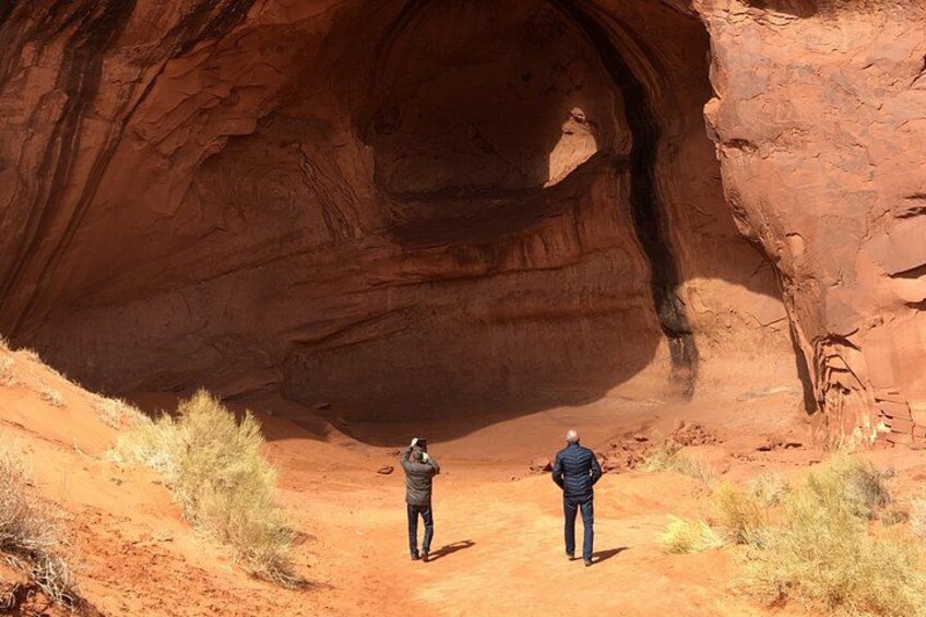 Deux personnes se tiennent sur un sentier de terre, devant une grande formation rocheuse rouge qui présente une ouverture ressemblant à une grotte. 3