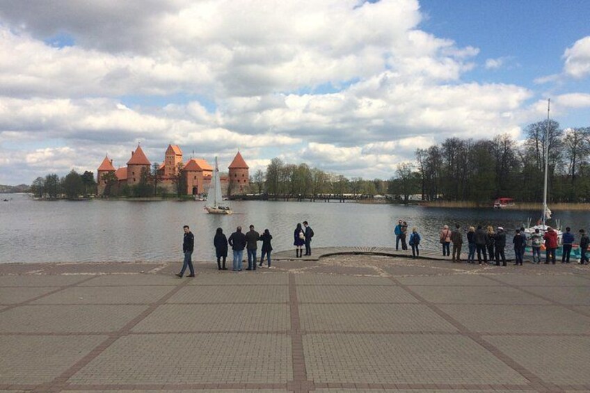 A riverside walkway with people, a castle, and a sailboat. 2