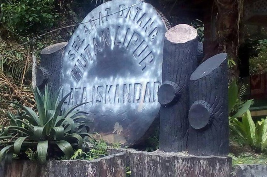 A metal sign with text, surrounded by trees and plants. 3