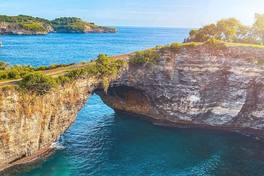 A coastal cliff with a natural arch formation, a clear blue sea, and distant green hills. 2