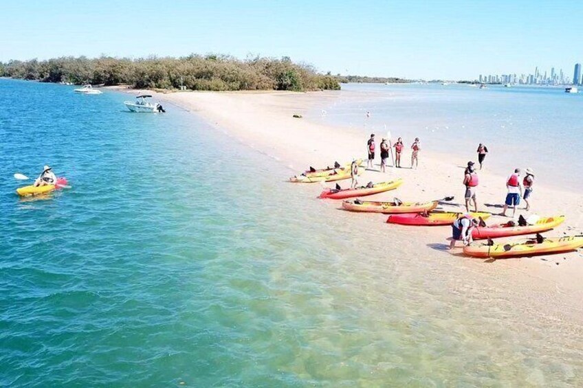 A beach with kayaks lined up on the shore, people standing and walking, and a clear blue sea. 1