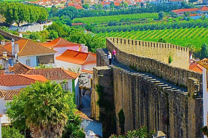 Un muro de piedra histórico con una escalera que lleva hacia él, rodeado de edificios con techos de tejas rojas y abundante vegetación. 2
