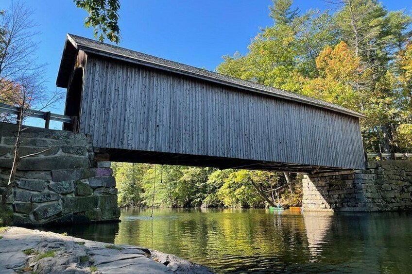 A wooden covered bridge over a calm river, surrounded by trees and stone walls. 4