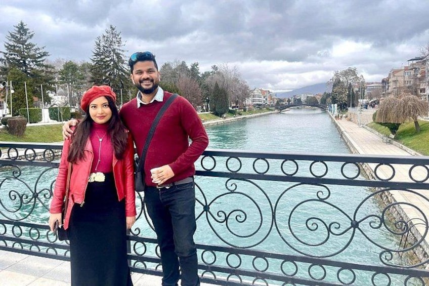 A couple standing by a river with a bridge in the background. 4