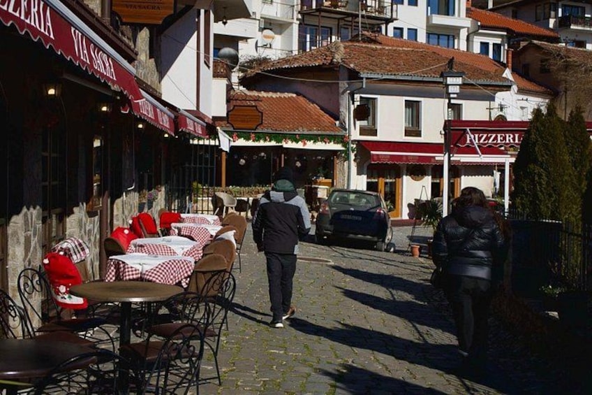A cobblestone street with outdoor seating at cafes, a parked car, and buildings with signage. 5