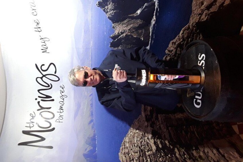 Una persona está de pie sobre un promontorio rocoso, sosteniendo un micrófono y una botella de cerveza Guinness, con vistas a un paisaje costero. 4