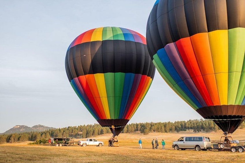 Twee luchtballonnen, waarvan één met kleurrijke strepen, en een voertuig met mensen en een aanhanger in een veld met bomen en heuvels op de achtergrond. 3
