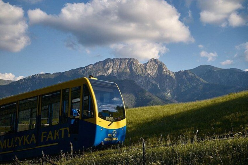 Un autobús amarillo y azul con la leyenda 'MUZIKA FAKTI' estacionado en una colina cubierta de hierba, con montañas al fondo. 5