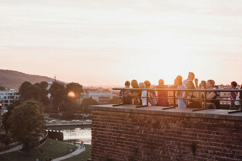 A group of people sitting on a rooftop terrace overlooking a river and cityscape during sunset. 4