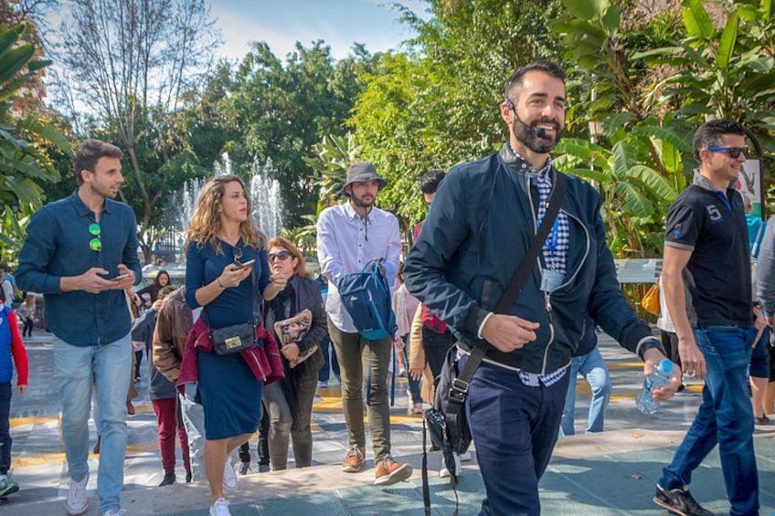 A group of people walking outdoors, some carrying bags, with trees and a fountain in the background. 2